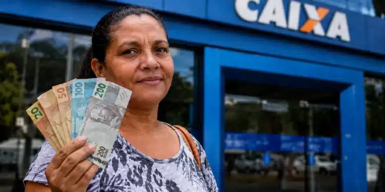 Mulher segura cédulas de real em frente a uma agência da Caixa, representando saque de R$ 2.900 disponível para CPFs com finais de 0 a 9.