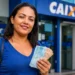 Mulher segurando cédulas de real em frente a uma agência da Caixa, representando saque de R$ 6.220 em cidades habilitadas.