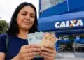Mulher segura cédulas de reais em frente a uma agência da Caixa, representando o saque de R$ 6.220 disponível até 1º de julho.
