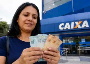 Mulher segura cédulas de reais em frente a uma agência da Caixa, representando o saque de R$ 6.220 disponível até 1º de julho.
