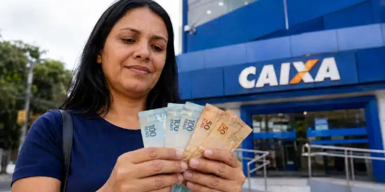 Mulher segura cédulas de reais em frente a uma agência da Caixa, representando o saque de R$ 6.220 disponível até 1º de julho.