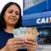 Mulher segura cédulas de reais em frente a uma agência da Caixa, representando o saque de R$ 6.220 disponível até 1º de julho.
