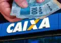 Mãos segurando cédulas de real em frente à fachada da Caixa Econômica Federal, ilustrando liberação de saque de R$ 6.220.