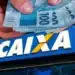 Mãos segurando cédulas de real em frente à fachada da Caixa Econômica Federal, ilustrando liberação de saque de R$ 6.220.