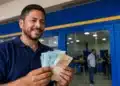 Homem sorridente segura cédulas de reais em agência bancária, representando a liberação do saque de R$ 6.220 pela Caixa.
