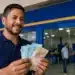 Homem sorridente segura cédulas de reais em agência bancária, representando a liberação do saque de R$ 6.220 pela Caixa.