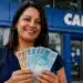 Mulher sorrindo com notas de reais em frente a uma agência da Caixa, representando o saque de R$ 6.220.