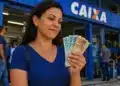 Mulher segura cédulas de real em frente a uma agência da Caixa, representando a solicitação do saque de R$ 6.220.