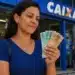 Mulher segura cédulas de real em frente a uma agência da Caixa, representando a solicitação do saque de R$ 6.220.
