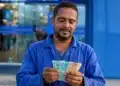 Trabalhador brasileiro conferindo notas de reais em frente a prédio bancário, representando o saque de R$ 6,2 mil do FGTS.