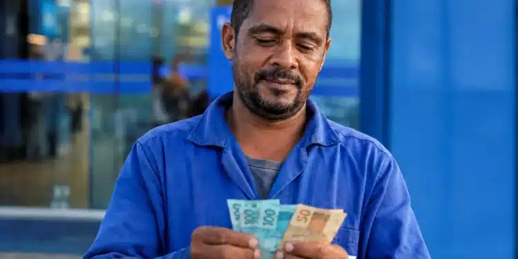 Trabalhador brasileiro conferindo notas de reais em frente a prédio bancário, representando o saque de R$ 6,2 mil do FGTS.