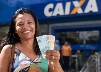 Mulher sorri segurando notas de real em frente a uma agência da Caixa, ilustrando saque de R$ 3.950 do FGTS até 30 de junho