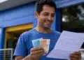 Homem brasileiro sorridente conferindo documento e dinheiro em frente a um banco, representando o saque de R$ 6.220.