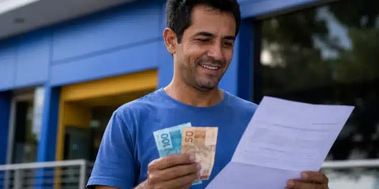 Homem brasileiro sorridente conferindo documento e dinheiro em frente a um banco, representando o saque de R$ 6.220.