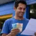 Homem brasileiro sorridente conferindo documento e dinheiro em frente a um banco, representando o saque de R$ 6.220.