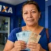 Mulher segura cédulas de real em frente a uma agência da Caixa, representando o saque de R$ 6,2 mil liberado para moradores de 8 cidades.