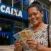 Mulher sorrindo ao segurar cédulas de real em frente a uma agência da Caixa, representando a solicitação do saque de R$ 6.220.