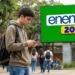 Estudante consulta celular em frente ao painel do Enem 2026, com destaque para a taxa de inscrição de R$ 85 e formas de pagamento.