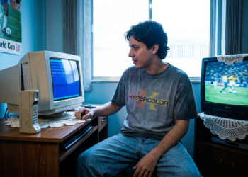 Jovem usa computador antigo de tubo ao lado de TV de tubo exibindo futebol