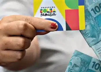 Mão segurando cartão do Bolsa Família com cédulas de real ao lado, representando os últimos pagamentos de junho do programa social.