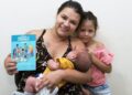 Mãe segura bebê no colo e exibe a Caderneta da Criança ao lado da filha em unidade de saúde