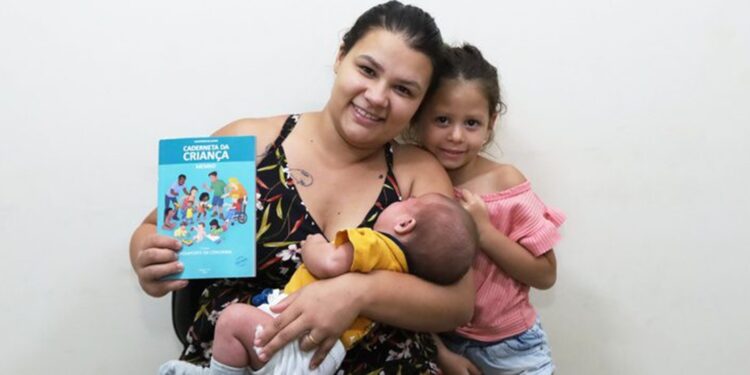 Mãe segura bebê no colo e exibe a Caderneta da Criança ao lado da filha em unidade de saúde