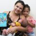 Mãe segura bebê no colo e exibe a Caderneta da Criança ao lado da filha em unidade de saúde