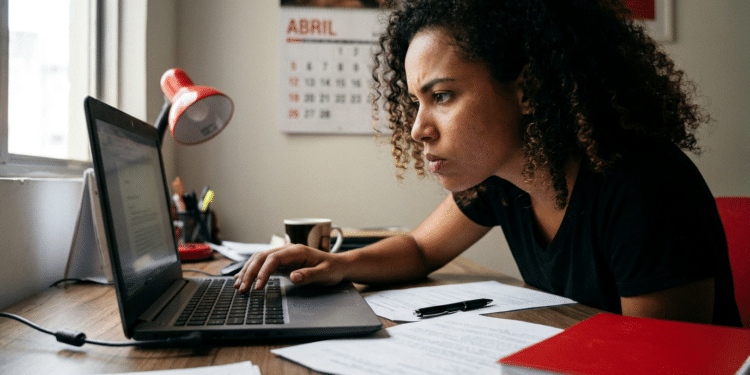 Mulher de cabelos cacheados usando camiseta preta trabalha em laptop sobre mesa com papéis, caneta e luminária vermelha.