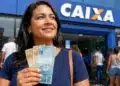Mulher segura cédulas de real em frente a uma agência da Caixa, representando a liberação ou solicitação do valor de R$ 2.900.