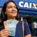 Mulher segura cédulas de real em frente a uma agência da Caixa, representando a liberação ou solicitação do valor de R$ 2.900.