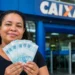 Mulher segura cédulas de R$ 100 em frente a uma agência da Caixa, representando o valor de R$ 6.220 disponível para CPFs de final 0 a 9.