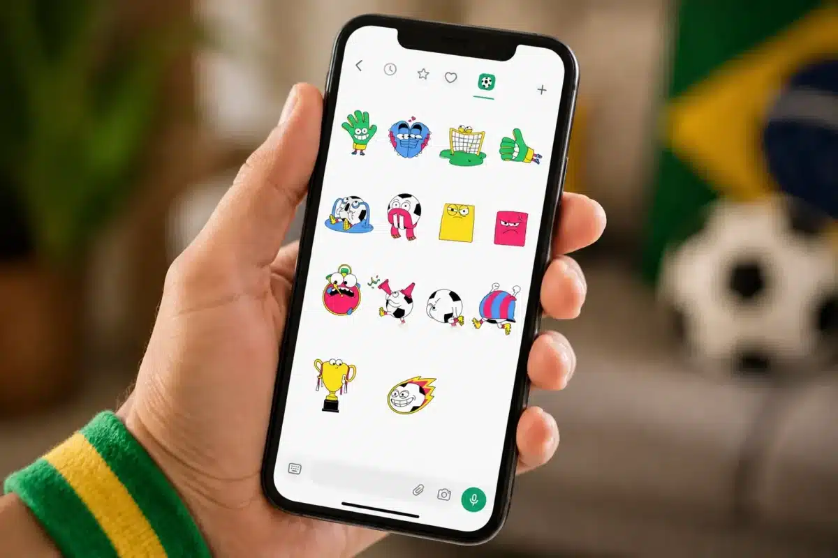 Mão segurando smartphone com figurinhas animadas da Copa do Mundo 2026 no WhatsApp, incluindo emojis de bola, chuteira, cartões e troféu.