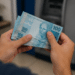 Mãos segurando três notas de 100 reais próximas a caixa eletrônico azul.