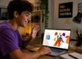 Jovem sorridente de camiseta roxa acena para notebook exibindo logo Rede Pode Falar, em mesa com livros.