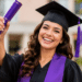 Jovem sorridente com beca preta e faixa roxa segura diploma durante cerimônia ao ar livre.