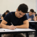 Jovem escreve concentrado em sala cheia de estudantes durante prova.