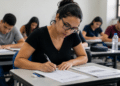 Mulher jovem de óculos preenchendo folha de respostas durante prova de concurso em sala com outros candidatos.