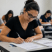 Mulher jovem de óculos preenchendo folha de respostas durante prova de concurso em sala com outros candidatos.