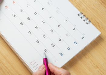 Mão segurando caneta rosa e calendário branco encima da mesa.