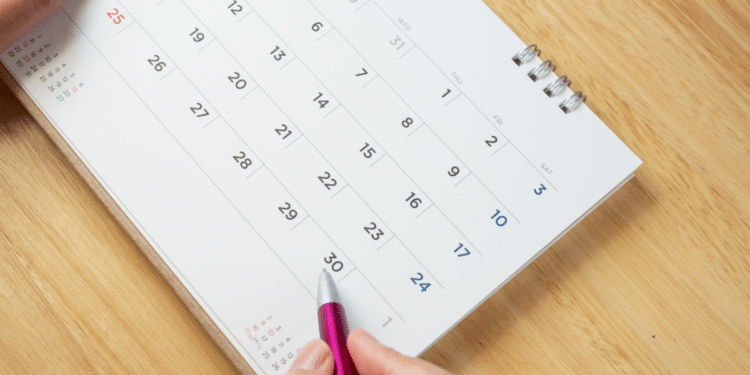 Mão segurando caneta rosa e calendário branco encima da mesa.