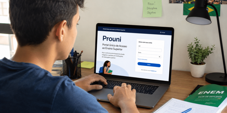 Rapaz acessa site do Prouni em notebook, caderno e guia ENEM sobre mesa, ambiente organizado.