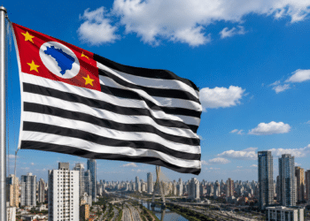 Bandeira de São Paulo tremulando ao vento com prédios altos ao fundo em dia ensolarado.