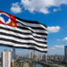 Bandeira de São Paulo tremulando ao vento com prédios altos ao fundo em dia ensolarado.