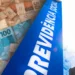 Notas de real ao lado de placa azul da Previdência Social com céu refletido.