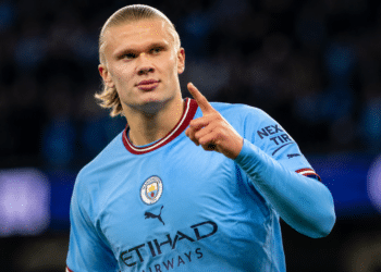 Jogador loiro do Manchester City aponta para o alto após marcar gol, uniforme azul claro e estádio ao fundo.
