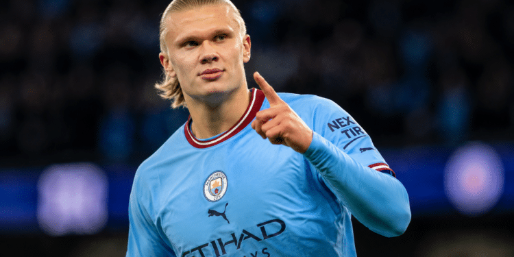 Jogador loiro do Manchester City aponta para o alto após marcar gol, uniforme azul claro e estádio ao fundo.