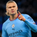 Jogador loiro do Manchester City aponta para o alto após marcar gol, uniforme azul claro e estádio ao fundo.