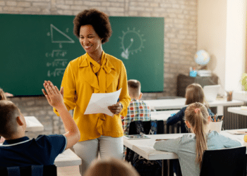 Professora entrega prova e sorri para aluno na sala com quadro verde.