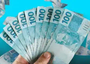 Mão segurando várias notas de 100 reais abertas em leque sobre fundo azul.