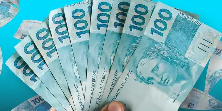 Mão segurando várias notas de 100 reais abertas em leque sobre fundo azul.